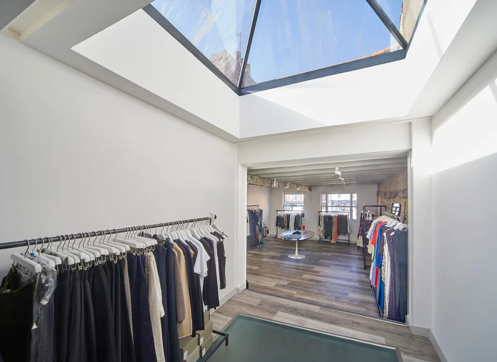 An interior view of a clothing store with clothes hanging on racks along the walls. The space has light-coloured wooden floors and white walls. A large glass skylight is visible in the ceiling, filling the room with natural light.