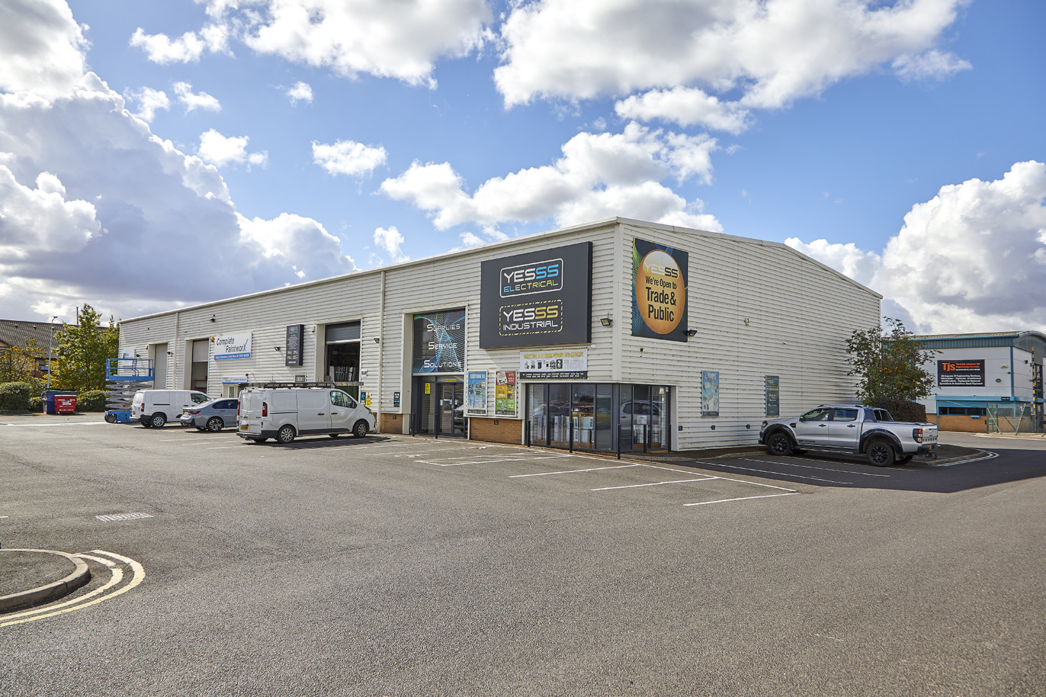 View from across a car park, of a white steel clad light industrial unit. Signage for Yesss Electrical on the building.