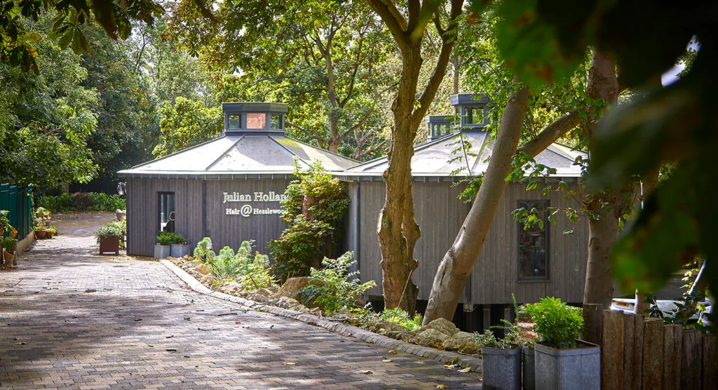 View, through trees, of 2 hexagonal shaped, grey wood-clad buildings.