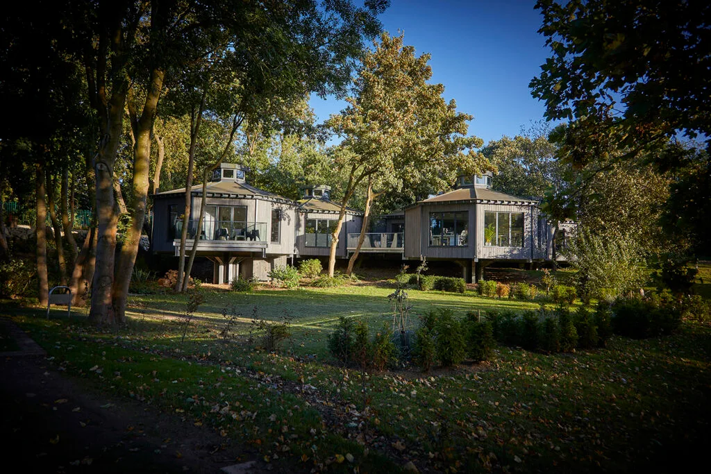 Grey wooden 'treetop' style building. 3 'pod' fronts showing glass balconies. surrounded by tress and grassland