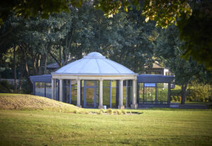 Rotunda shaped building with full glas wall windows and steel framed square extensions to each side, set in parkland