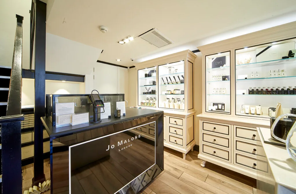 A retail counter inside a Jo Malone store. The counter is sleek and black with "Jo Malone London" printed on the front. Behind the counter, there are two tall, white display cases with glass shelves holding various products. The floor is light-colored wood.