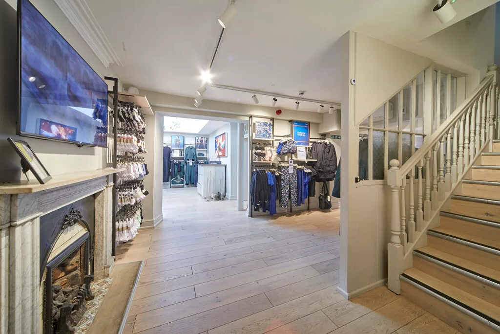 An interior view of a retail space with light-colored wooden floors. A staircase with a wooden banister and a large fireplace are visible in the foreground. Clothing displays and racks are visible further into the shop. A television screen is mounted above the fireplace on the left.