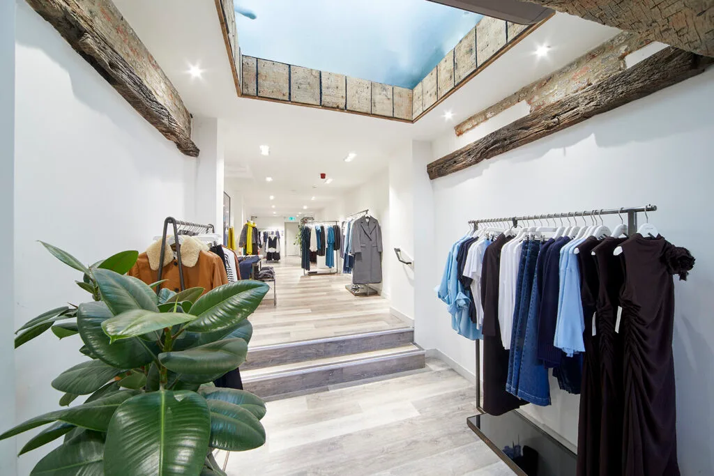 An interior view of a clothing store with clothes racks and wooden floors. A skylight is visible in the ceiling, with old wooden beams framing the opening. A set of stairs leads down to a lower level with more clothing displays. A large green plant is in the foreground.