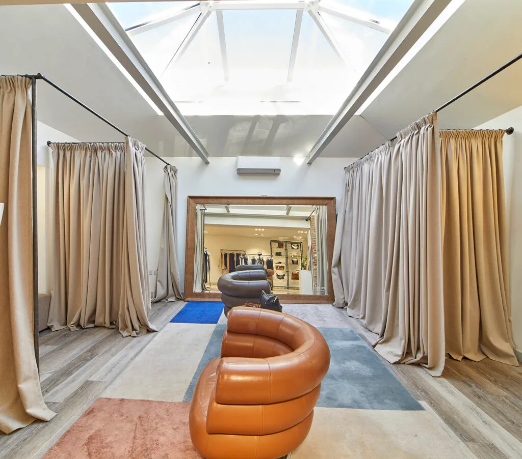 A dressing room area with two large, curved leather chairs, one brown and one gray, sitting on a colorful geometric rug. The space is partitioned by light-colored curtains hanging from black rods on both sides. A large mirror in a wooden frame stands against the back wall, reflecting the room. The ceiling has a prominent glass skylight, letting in a lot of natural light.