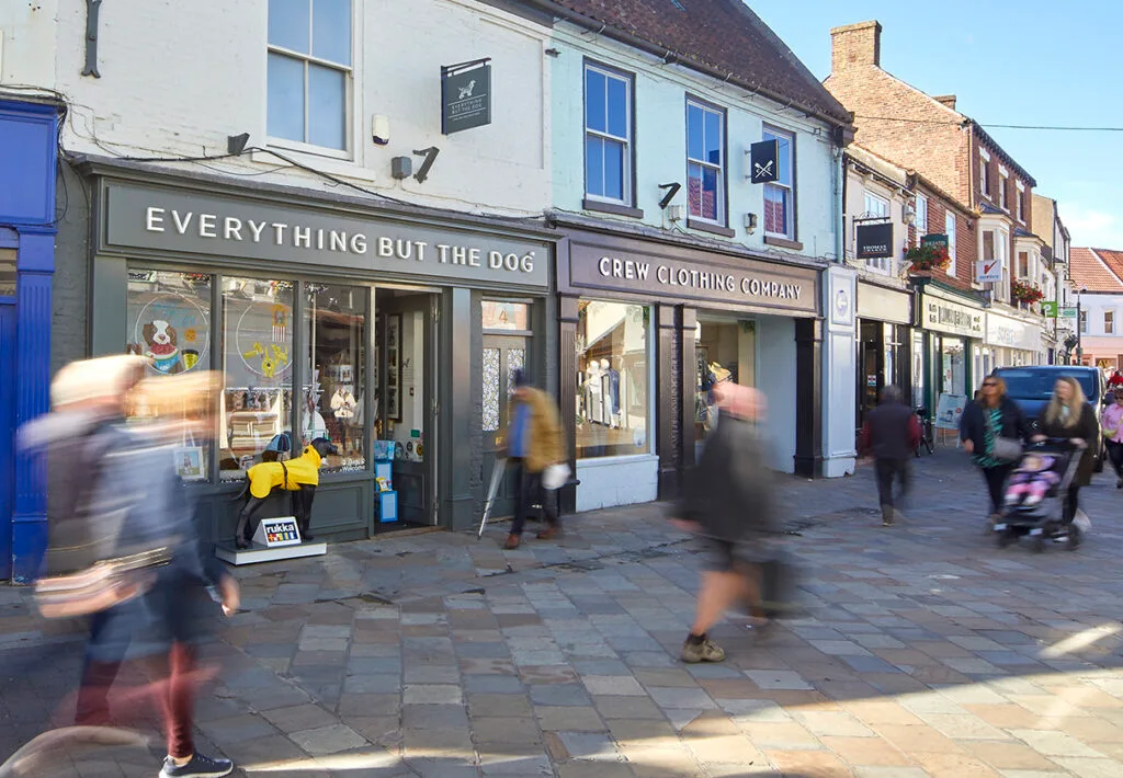 A blurred, street-level view of a bustling town square. Several storefronts are visible, including "Everything But the Dog" and "Crew Clothing Company." People are walking along the cobblestone pavement in a blur of motion. The buildings are a mix of brick and painted exteriors.