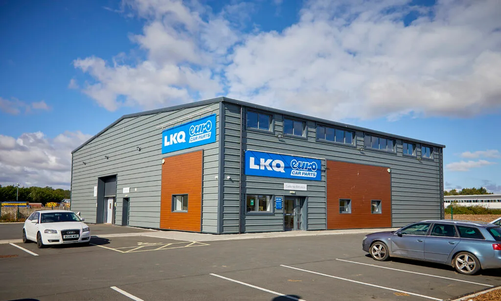 A two-story grey and brown industrial building with a large sign that reads "LKQ Euro Car Parts." The building has a roll-up garage door and several windows. Two cars are parked in the lot in the foreground. The sky is blue with scattered white clouds.