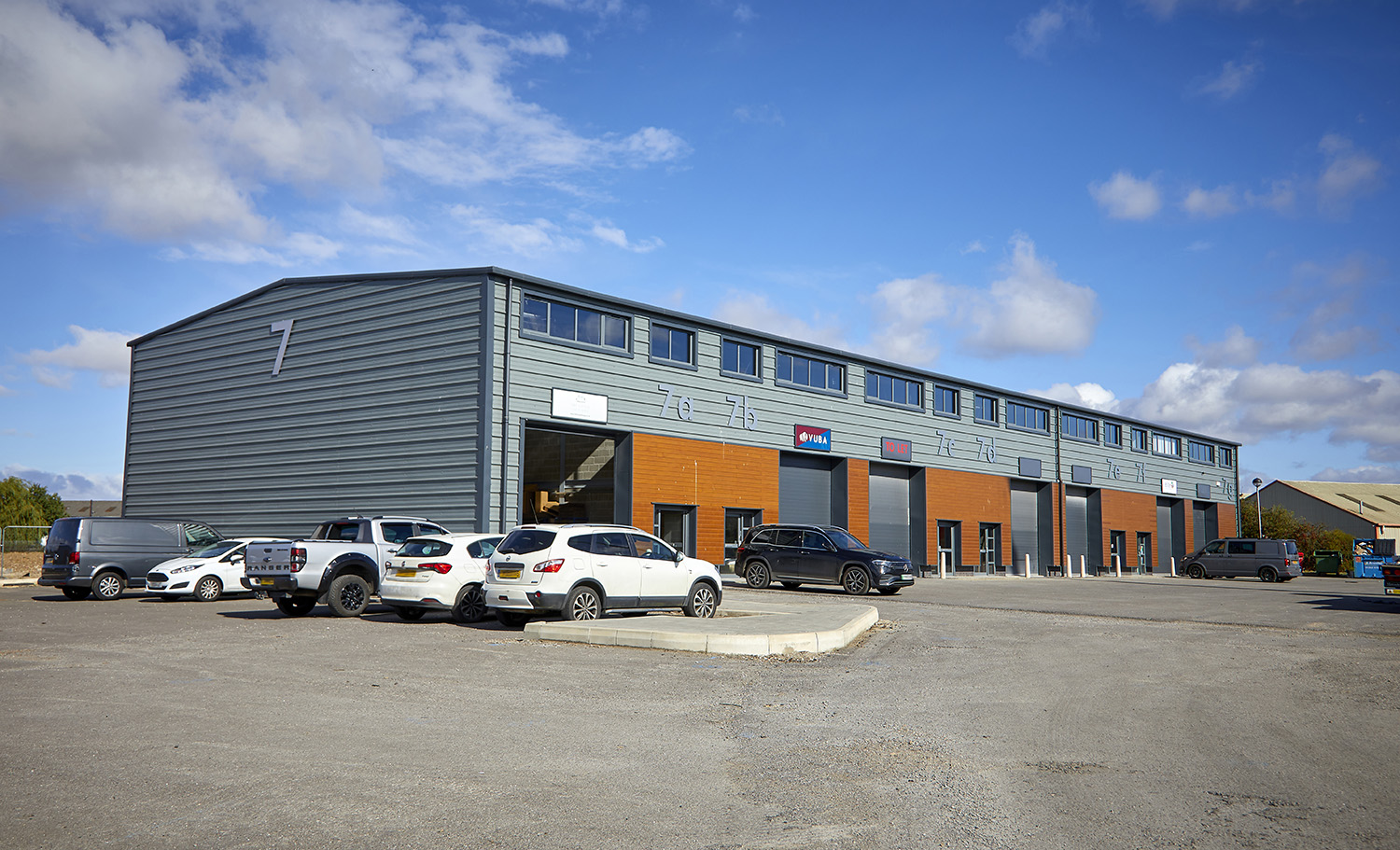 Side end view of trade units at Capital Park Beverley. The units are grey steel and brown wood cladded. The image shows carpark and several parked cars