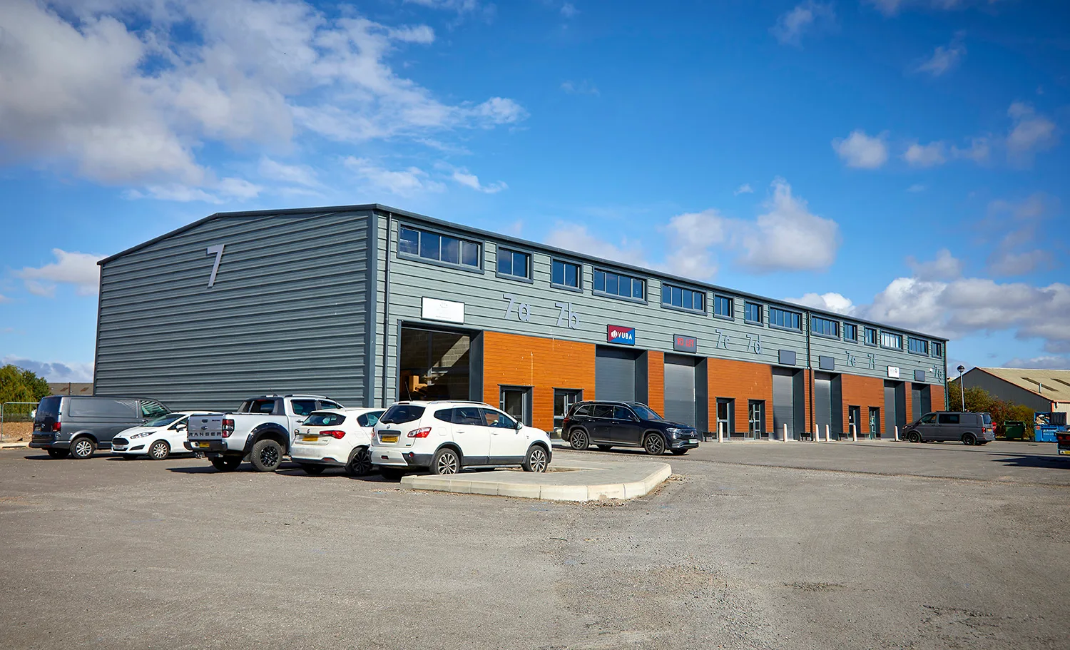 Side end view of trade units at Capital Park Beverley. The units are grey steel and brown wood cladded. The image shows carpark and several parked cars