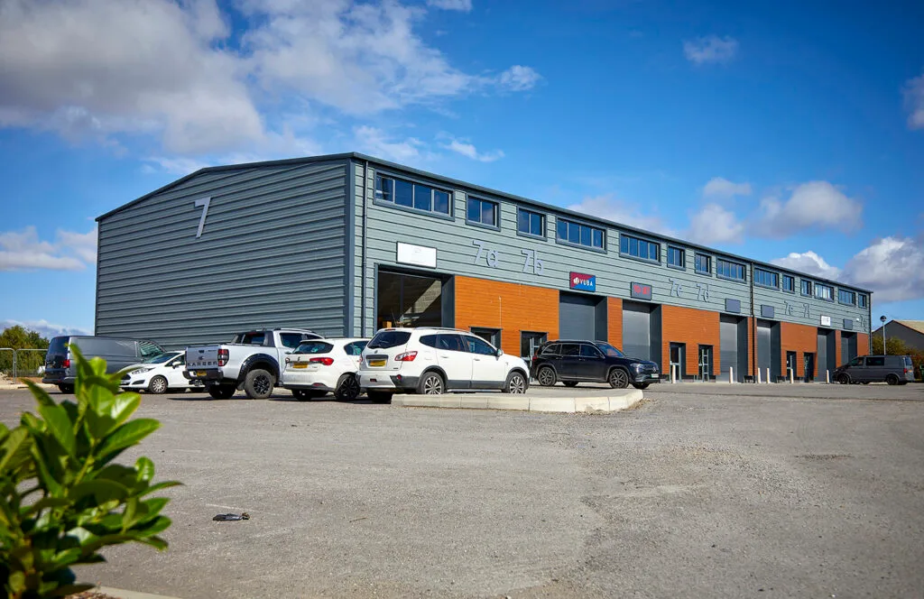 A wide-angle view of a long, gray industrial building with orange and black doors. The building has a sign with the number 7 on the left and a row of windows across the top. Several vehicles are parked in the lot in front of the building. The words "Capital Park, Beverley" are written in large white letters across the center of the image.