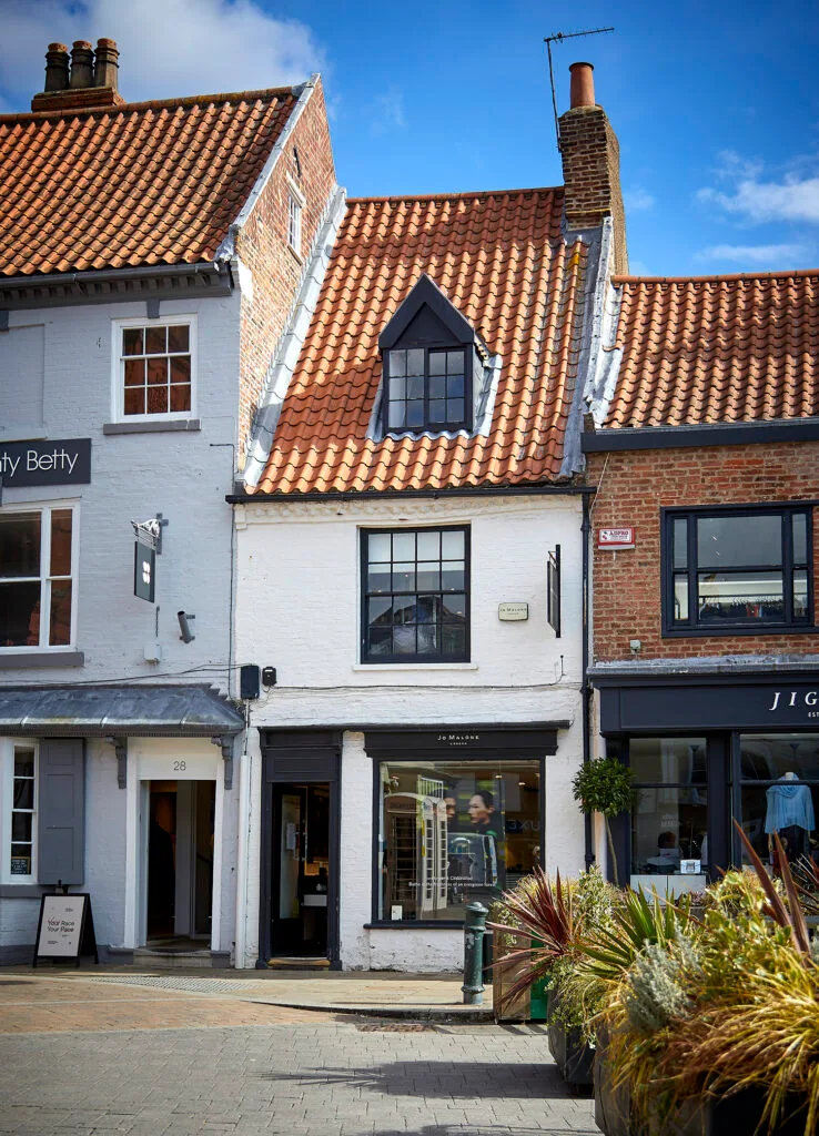 A white, narrow, two-story building with a red tile roof and a prominent dormer window. The building is a storefront with large windows and a sign above the door. It is located on a cobblestone street, nestled between a grey building and a brick building, both with shops. There are green plants in pots in the foreground.