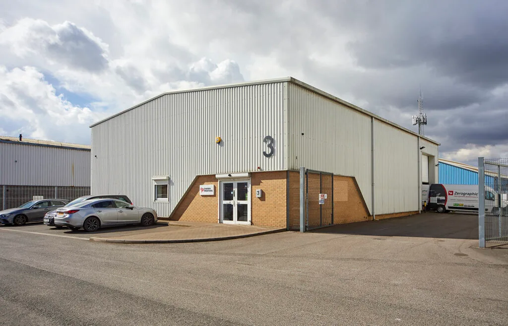 A large, white industrial building with a small brown brick section around the entrance. A large number 3 is on the front of the building. Two cars are parked in front of the building, and a chain-link fence is on the right. The sky is blue with large, white clouds.