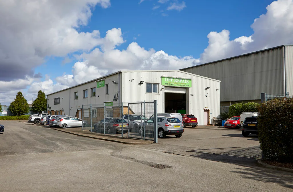 Exterior view of a white industrial unit with a car park in front. A sign above the main door reads "DIY Repair". There are several cars parked in the foreground.