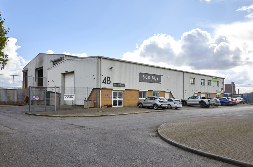 Side view of a white steel and brick clad industrial building. There is a gated compound to the rear of the building and several vehicles parked to the side