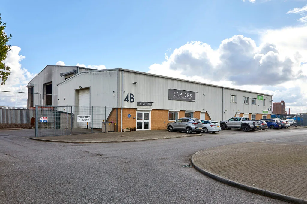 A grey industrial building with a white sign that says "Scribes." The address "4B" is written on the front of the building. Several cars are parked in the lot, and a chain-link fence is to the left. The sky is blue with scattered white clouds.