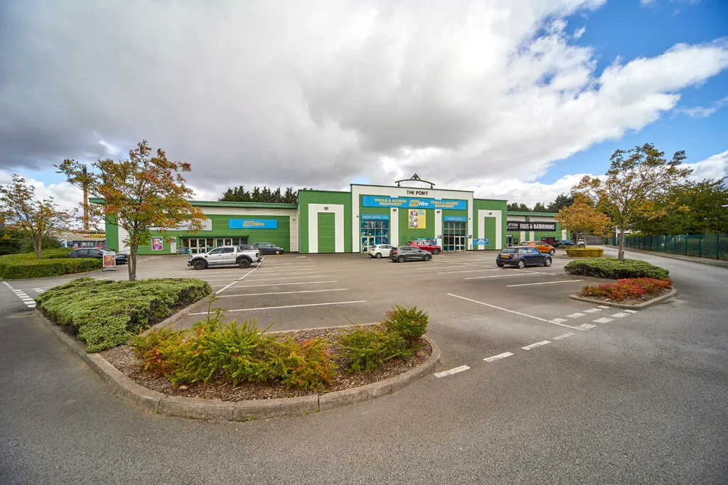 An industrial park with a large green building in the centre. The central sign shows the words 'THE POINT'. The lot in front of the building is a paved parking area with several cars and some landscaped islands with low-lying plants.