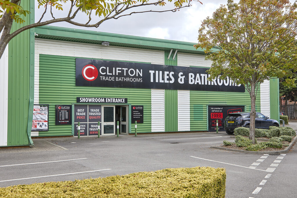 View from across a car park, of a green steel clad light industrial unit. Signage for Clifton Bathrooms on the building.