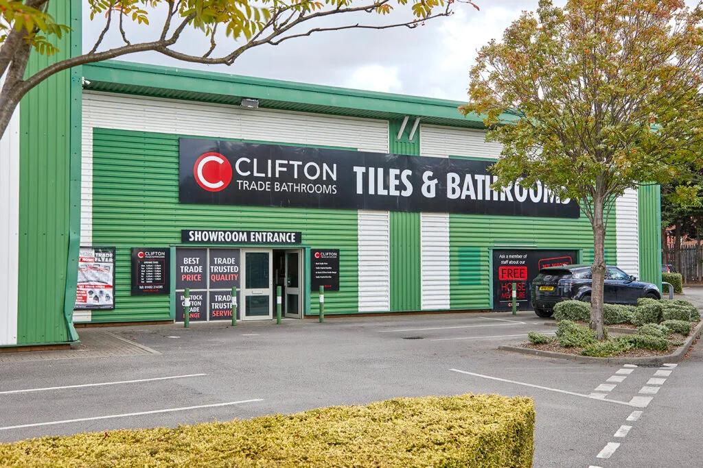 A wide, one-story commercial building with green and white striped siding. The building has a sign that reads "Clifton Tiles & Bathrooms" in black lettering against a green background, with a red "C" logo. There is a "Showroom Entrance" sign above the double doors. A car is parked to the right of the entrance.
