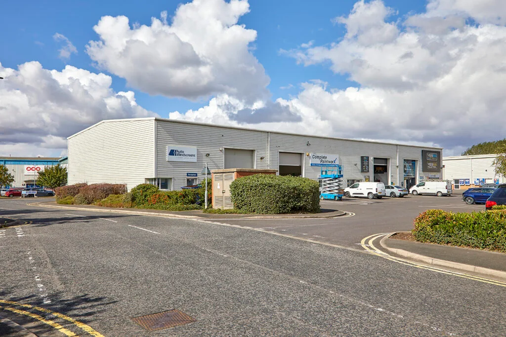 A long, grey industrial building with a red and black logo on the front. A large roller door is in the centre of the building, and a few cars are parked in the lot in front of it. There are green shrubs and trees in the foreground and a blue sky with white clouds.