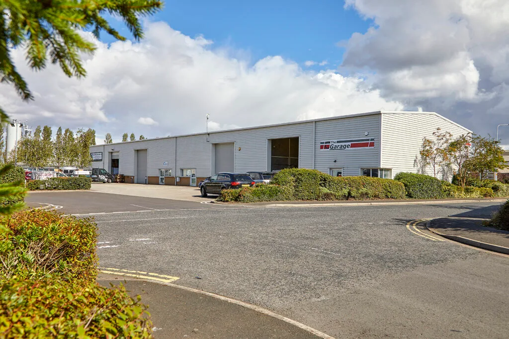 A long, grey industrial building with a red and black logo on the front. A large roller door is in the centre of the building, and a few cars are parked in the lot in front of it. There are green shrubs and trees in the foreground and a blue sky with white clouds.