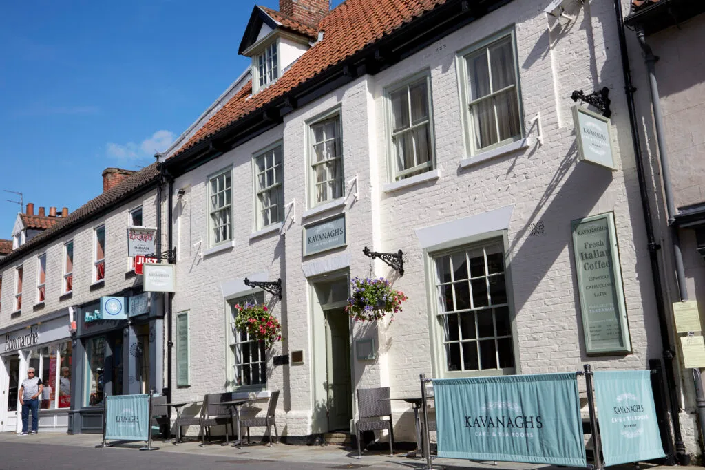 A multi-story white brick building with light green window frames. A sign over the door reads "Kavanagh's," and another sign with the same name is on a barrier in the foreground. Hanging baskets with red and pink flowers are on either side of the entrance. Two tables and chairs are set up outside on the pavement.