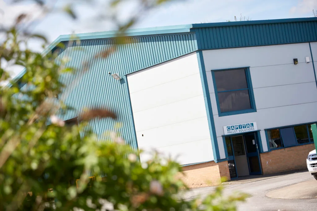 A light blue and white industrial building with a small sign above and entrance door. The building has a large white panel on the front and is partially obscured by out-of-focus green foliage in the foreground.