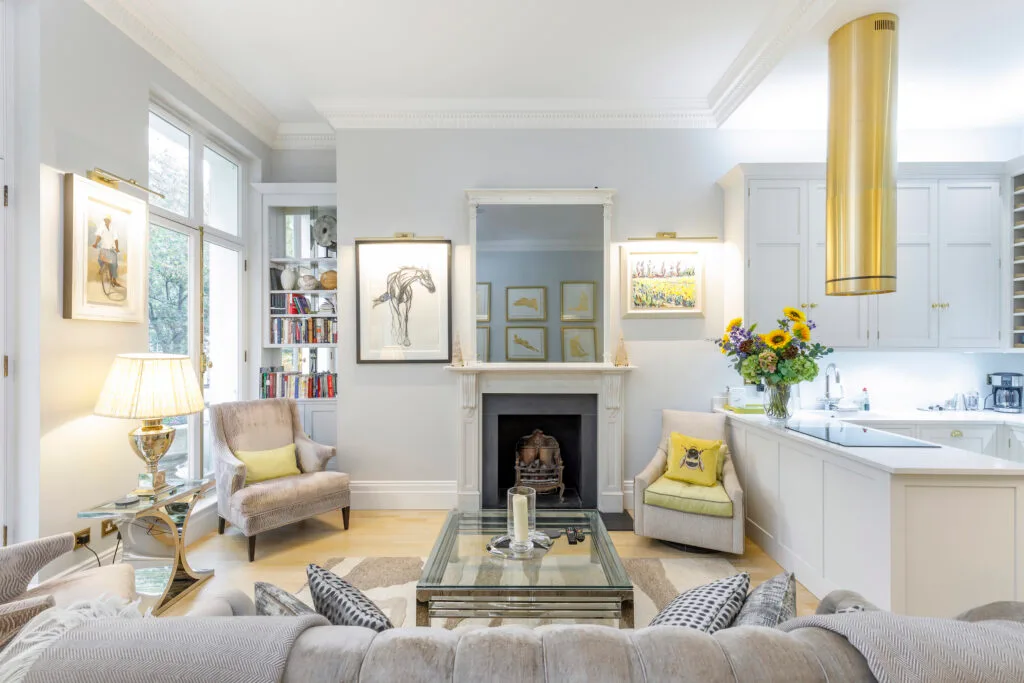 Residential Portfolio: Brooklands Property Holdings A brightly lit living room in a residential property, with a fireplace and an adjoining kitchen. The room features a light color palette with a gray fireplace, two upholstered armchairs, and a glass coffee table. Artwork hangs on the walls, and a large mirror is mounted above the fireplace. The kitchen area has white cabinets and a prominent gold extractor fan over the hob. A vase of sunflowers sits on the kitchen counter.