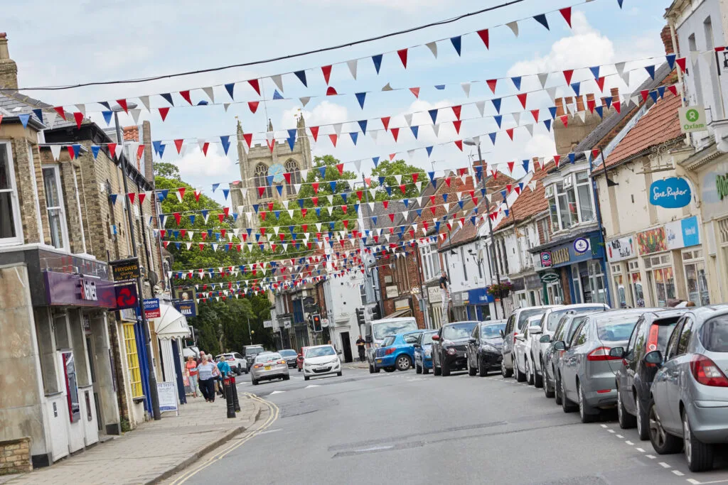 A view down a busy street in a town, with red, white, and blue bunting strung across the road. The street is lined with cars and several two-story buildings housing various shops and businesses. A church steeple is visible in the background, and the sky is partly cloudy.