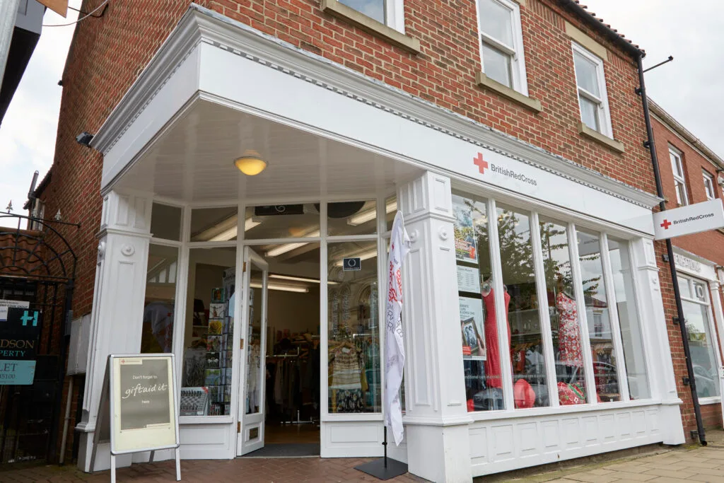 A brick building with a white storefront and large windows. The sign for the shop reads "British Red Cross." The windows display various items of clothing, and the front door is open. The top half of the building is red brick with a large window and a smaller window.