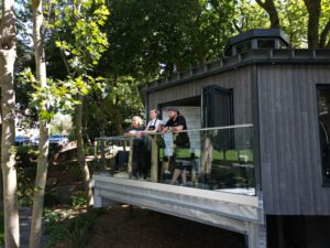 Grey wooden 'treetop' style building. Showing glass balcony with 3 people stood on it, chatting.