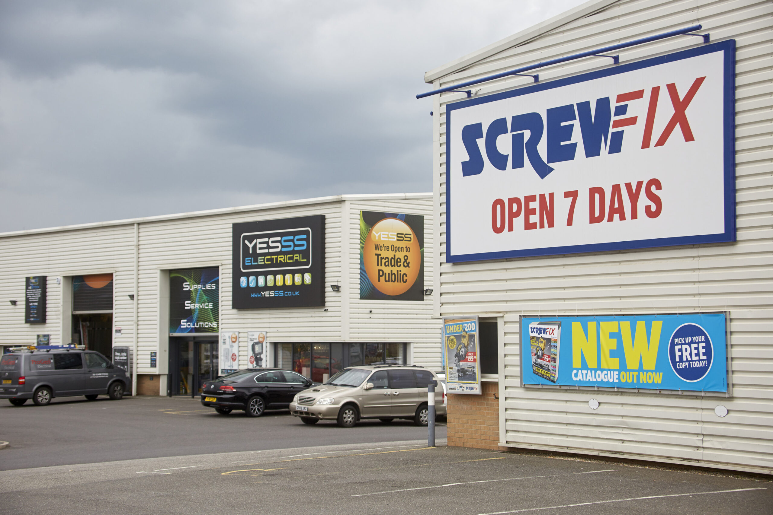 Partial view of 2 white-clad, trade units. The foremost shows Screwfix signage and the one at distance shows Yesss Electrical signage