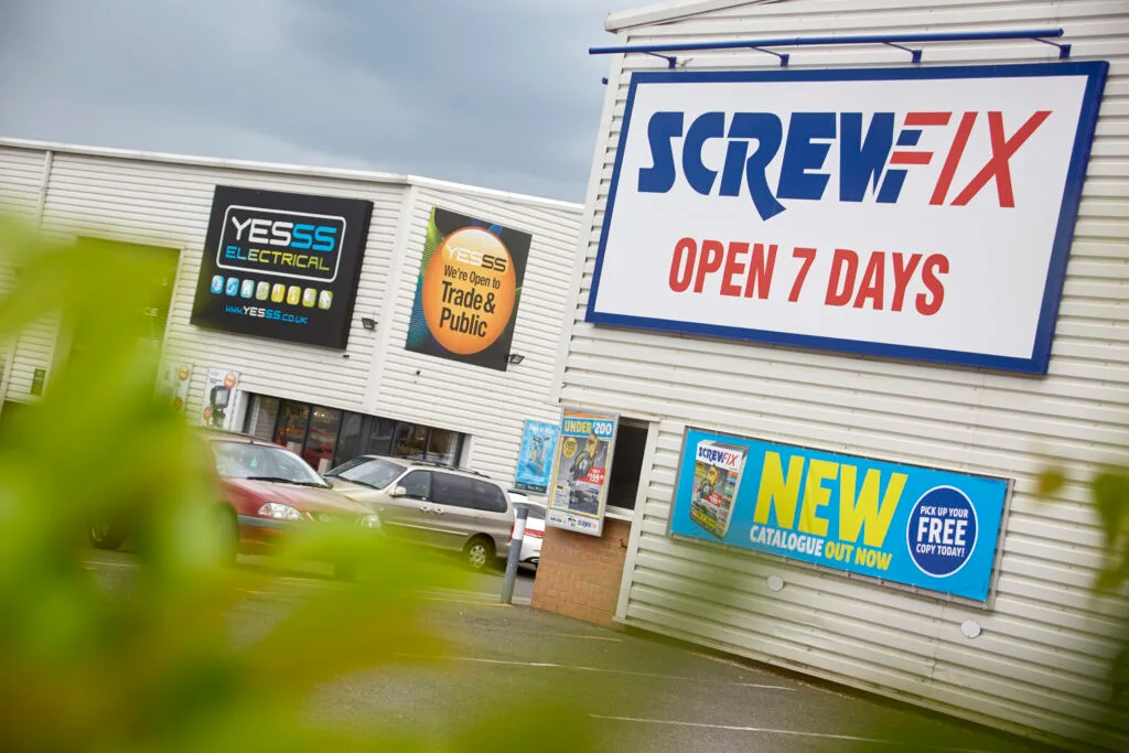 A large, white commercial building with signs for "Screwfix" and "YESSS Electrical." A large blue and red sign for Screwfix announces they are "OPEN 7 DAYS." Another sign for YESSS Electrical reads "We're Open to Trade & Public." The building is partially obscured by out-of-focus green plants in the foreground. Several cars are parked in the lot.