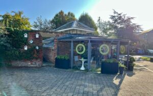 View across a carpark of a conservatory style structure leading from a brick built building, This is a deli at Hesslewood Office Park