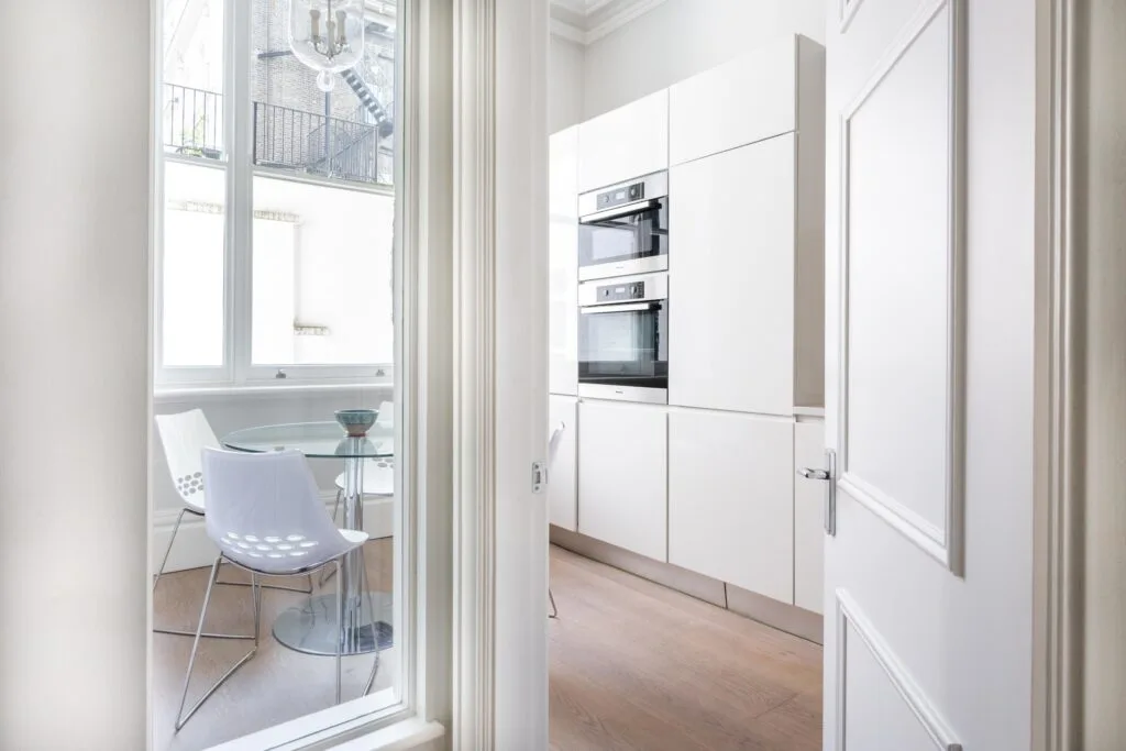 Residential Portfolio: Brooklands Property Holdings A clean, modern kitchen with white, handleless cabinets. A pair of built-in ovens is visible on the wall. To the left, a large window looks out onto a dining area with a glass table and a white chair.