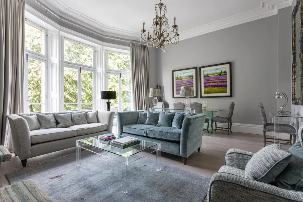 Residential Portfolio: Brooklands Property Holdings A sophisticated living room with tall, curving bay windows that let in natural light. The room is decorated in a muted gray and blue color scheme, featuring a gray sofa, a blue velvet sofa, and a matching armchair. A glass coffee table sits on a large gray rug, and two framed pictures of lavender fields hang on the wall. A small dining table and chairs are in the background.