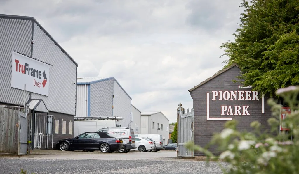 A wide-angle, low-elevation shot of an industrial park with a large grey building on the left and a building with a red sign on the right. The sign on the right building has the words "PIONEER PARK" in a large, white font. The foreground is a paved parking area with some vans and cars. The words "Pioneer Park, Hull" are overlaid in large white letters across the centre of the image. The sky is grey and cloudy.