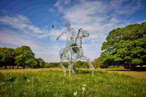 Wire-framed statue of man playing polo on a horse. The statue is set in grassed parkland, with wildflowers and trees surrounding