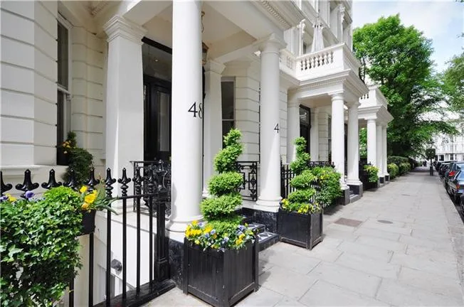 Residential Portfolio: Brooklands Property Holdings A close-up view of the front of a classic white terraced house. Two large white columns flank the entrance, with the number 48 visible on the column to the left. A black planter box with a manicured spiral topiary and colorful flowers sits in front of the doorway. A black metal railing lines the short staircase.