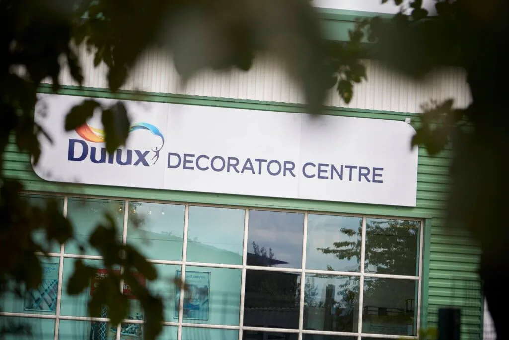 A close-up view of the front of a "Dulux Decorator Centre." The sign is large and white with the Dulux logo and the words "DECORATOR CENTRE" in large blue letters. The lower part of the image shows the shop's glass-fronted windows.