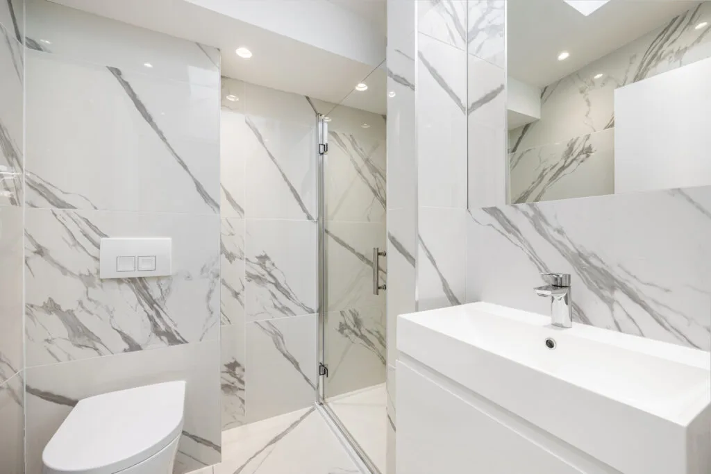 Residential Portfolio: Brooklands Property Holdings A bright, modern bathroom with white marble-effect tiles that have prominent gray veining. A glass shower door is visible in the center. A white sink vanity is on the right, and the side of a toilet is on the left.