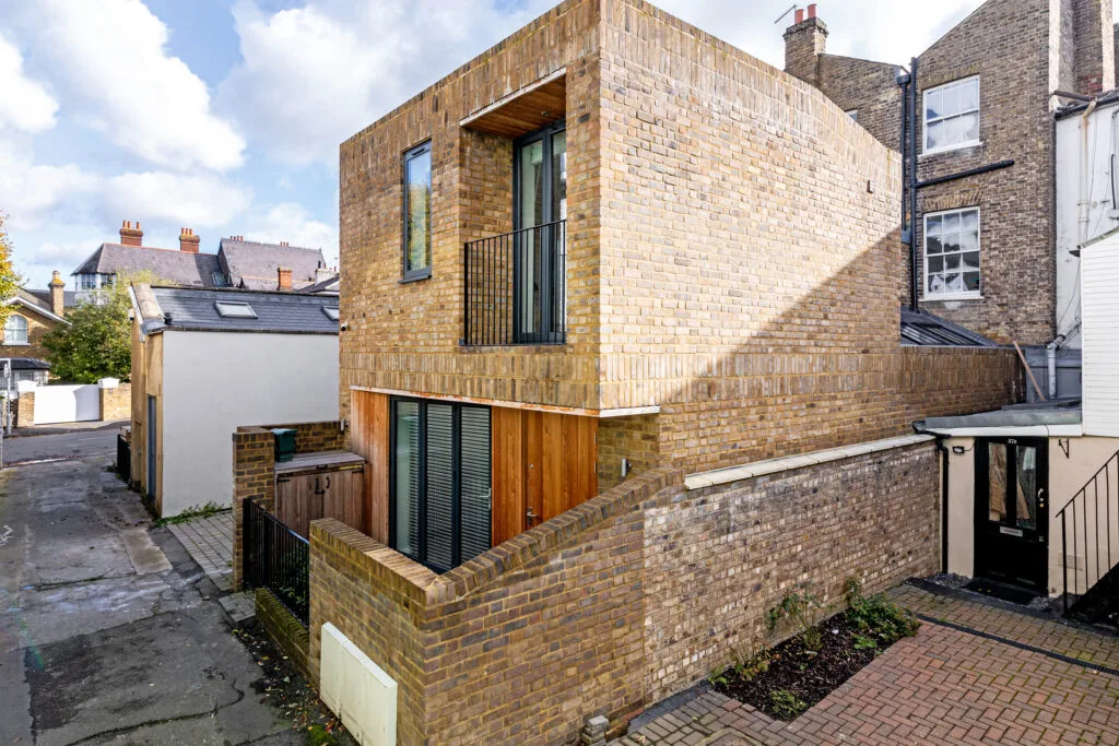 Residential Portfolio: Brooklands Property Holdings A modern, multi-story building made of yellowish-brown brick. The building features a small balcony with a black railing on the second floor. A wooden facade surrounds the ground-floor entrance, and a brick wall encloses a small patio area