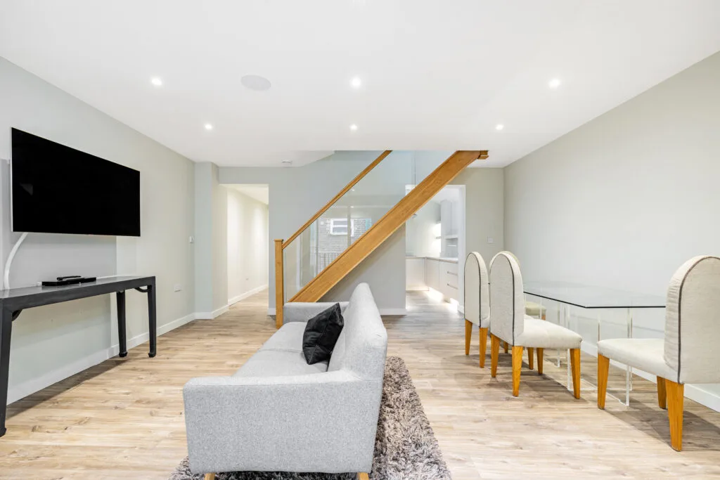 Residential Portfolio: Brooklands Property Holdings A clean, modern open-plan living and dining area. A light gray sofa is in the foreground, facing a wall-mounted television. To the right, a clear glass dining table is set with four chairs. A wooden staircase with a glass railing leads to the second floor, and the kitchen is visible in the background.