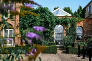 Partial view of a brick built building with flowers and greenery growing on the walls. The centre of the image shows a conservatory style roof.