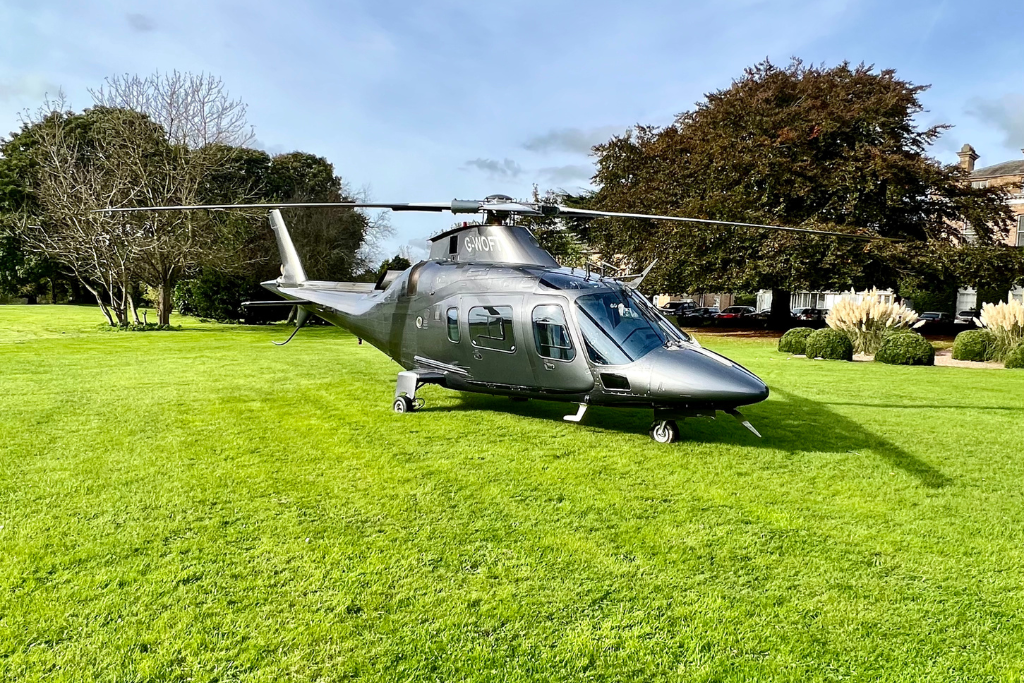 Grey helicopter, landing on grassed parkland. Trees to background