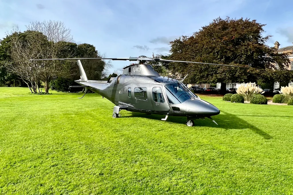 A side-view shot of a modern, dark grey helicopter with a glossy finish, parked on a manicured green lawn. The helicopter's rotor blades are stationary, and its landing gear is on the grass. In the background, there is a large building, a few trees, and a low hedge. The sky is partly cloudy.