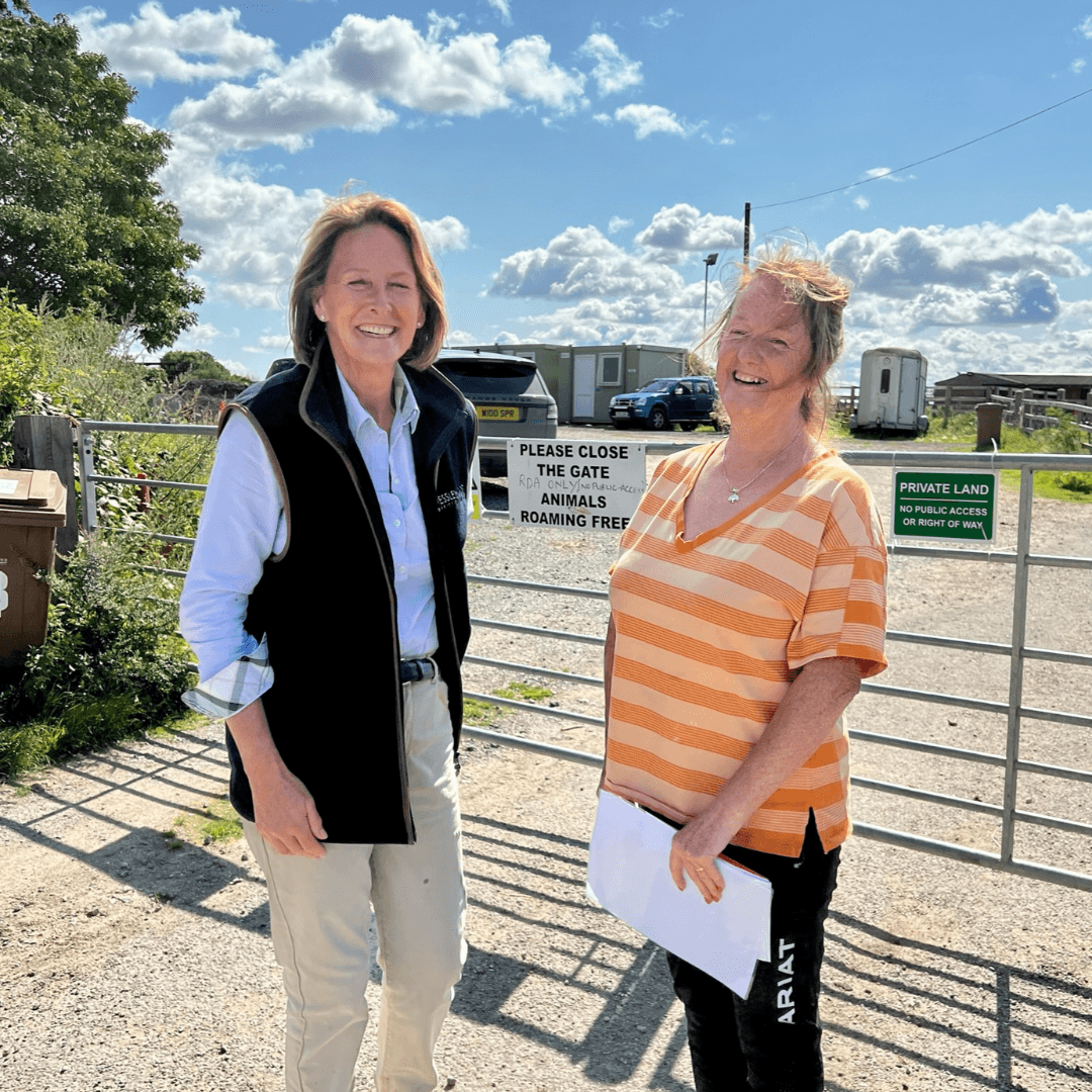 2 women stood next to each other, with horse compound in background. Both women smiling toward the camera