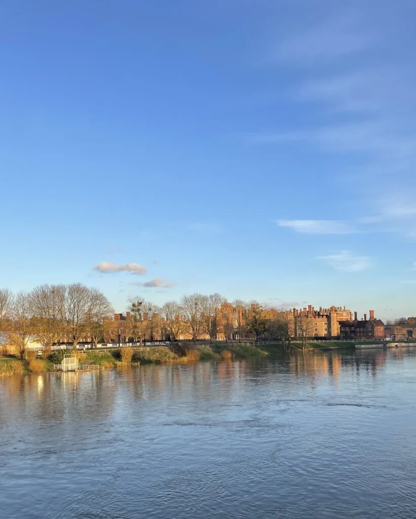 Hampton Court A view across a wide, calm river towards a tree-lined bank. Behind the bare trees, a large, historic brick building with many turrets and chimneys stretches along the waterfront. The sky above is clear and blue with a few small clouds.
