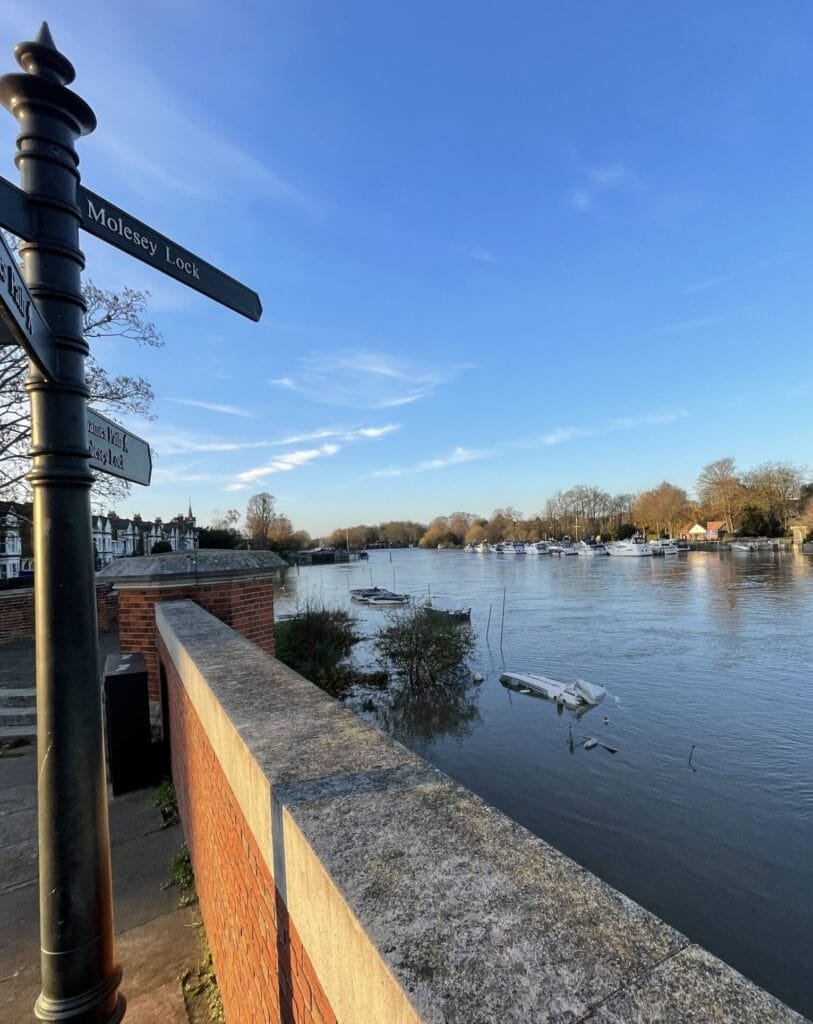 Mosely Lock A scenic view of a wide river with high water levels, almost reaching the top of the brick wall in the foreground. Several small boats are moored along the opposite bank, and a signpost on the left points towards "Molesey Lock." The sky is clear and blue.