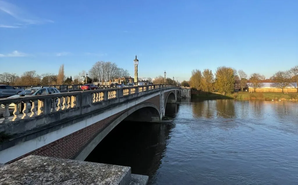 East Mosely A bridge made of brick and stone with an ornate railing, spanning a river. Cars are driving across the bridge. A lamppost with a globe light is visible on the bridge. The sky is bright blue, and the sun is low, casting a warm light on the bridge. Bare trees line the riverbank on the far side.
