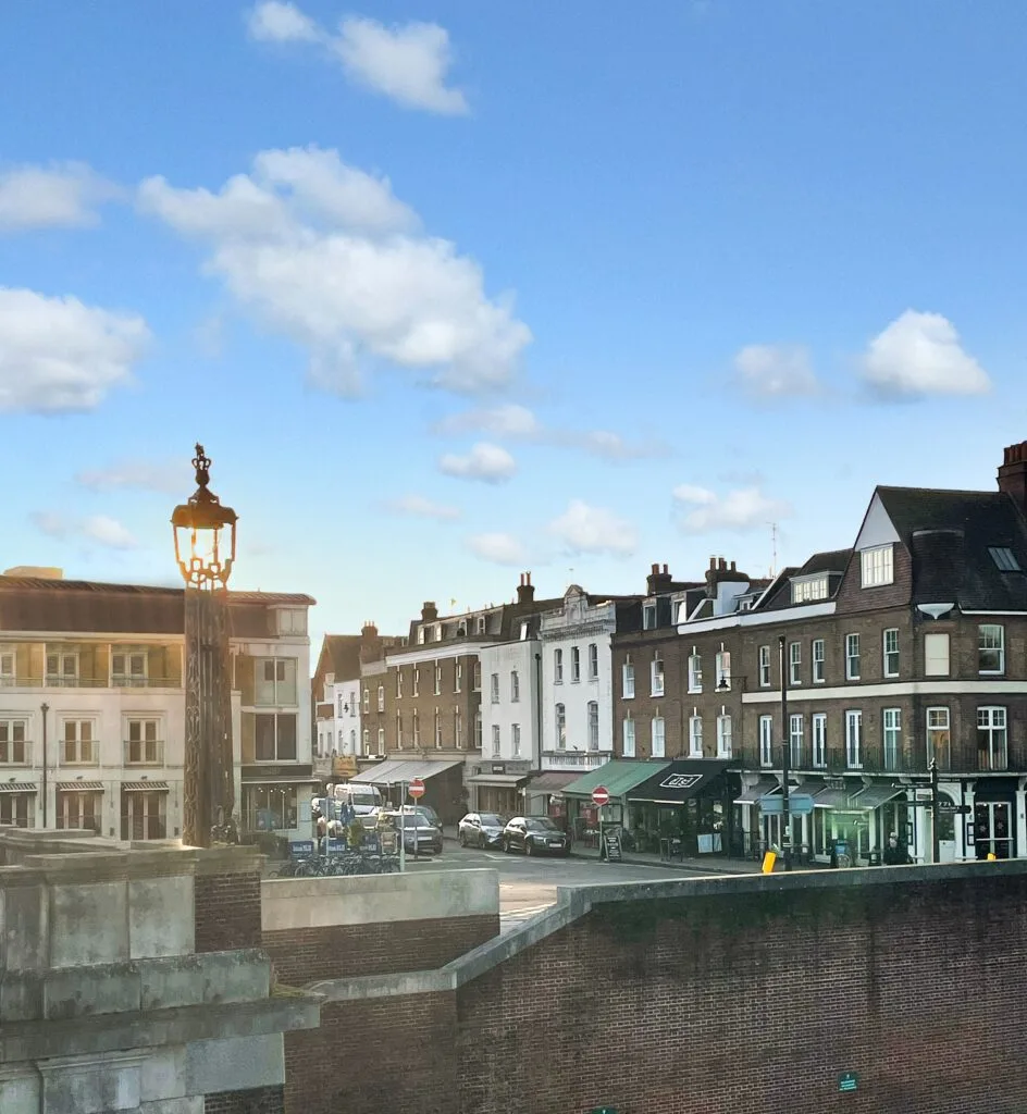 A low-angle view of a brick wall and a large, ornate street lamp in the foreground. In the background, a street is lined with several brick and white buildings, which house shops and businesses. The street is busy with people and cars. The sky is blue with scattered white clouds.